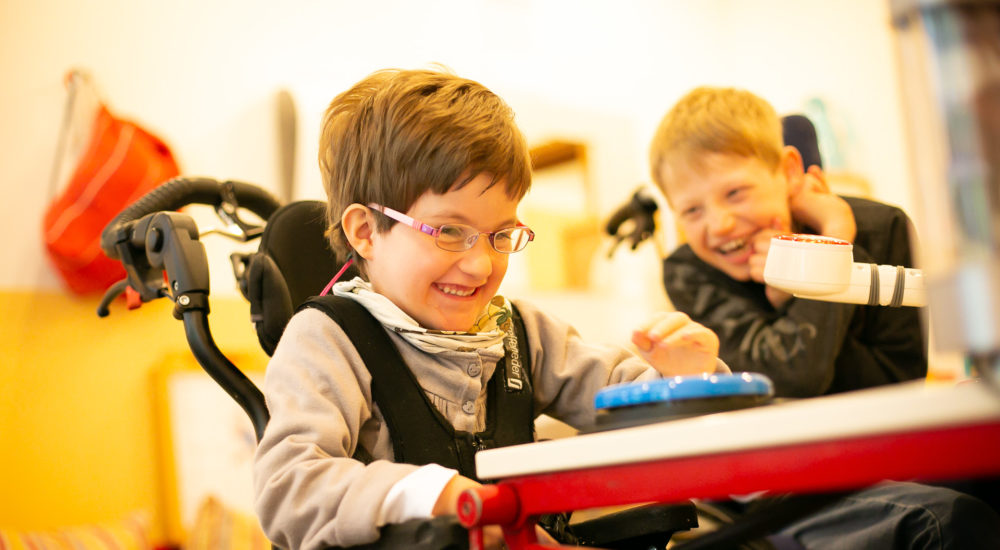 Eine Schülerin im Rollstuhl sitzt am Tisch. Ein weiter Schüler lacht sie an. ©Helfende Hände/Fabian Helmich
