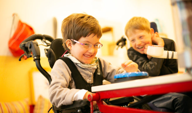 Eine Schülerin im Rollstuhl sitzt am Tisch. Ein weiter Schüler lacht sie an. ©Helfende Hände/Fabian Helmich